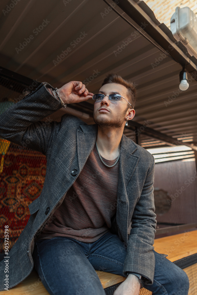 Handsome hipster model man with trendy coat, sweatshirt and jeans wears a blue sunglasses and sits on the street