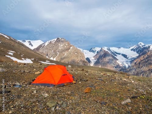 Summer camping in mountain. Bright alpine landscape with vivid orange tent at very high altitude with view to high mountain and large glacier in dark clouds. Awesome mountain scenery with tent.