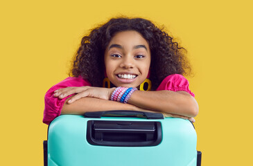 Studio shot of happy child with suitcase. Portrait of cheerful kid with travel bag. Beautiful African American girl with curly hair in trendy outfit with vacation baggage isolated on yellow background