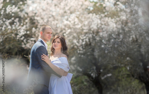 a guy in a suit and a girl in a white dress are standing in a garden with flowering trees and hugging. © Olga