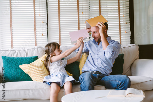 Happy father and cute daughter sitting on sofa and holding book on heads and looking each other. Smiling single father playing wiht little girl enjoying time at home.