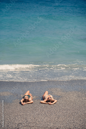 Fototapeta Naklejka Na Ścianę i Meble -  Two girls girlfriend are sitting on the sea sandy shore and the waves soaked them in bathing suits on a sunny warm day