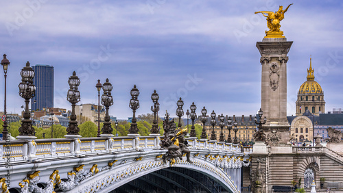 The ornate Pont Alexandre III in the downtown of Paris, France with the golden dome of the Invalides and the Tour Montparnasse in the background below an overcast sky photo