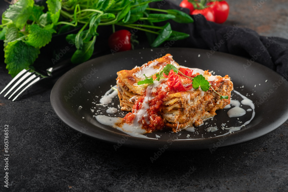 A portion of meat lasagna with bolognese sauce and mozzarella cheese on a black plate. Italian dish with layers of pasta and minced beef A portion of meat lasagna with bolognese sauce and mozzarella cheese on a black plate. Italian dish with layers of pasta and minced beef