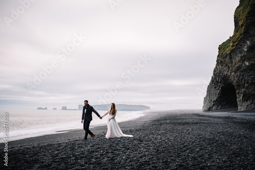 Bride and groom in wedding outfits walk on the Black Sand beach with the view to the Atlantic ocean and horizon. Epic view in Iceland Elopement.
 photo