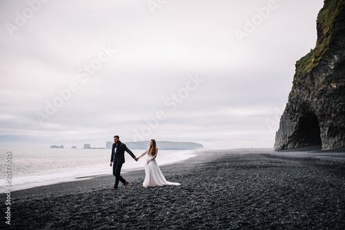 Bride and groom in wedding outfits walk on the Black Sand beach with the view to the Atlantic ocean and horizon. Epic view in Iceland Elopement.
