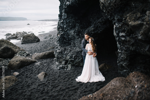 Bride in white silk dress and groom in jacket kiss each other in a basalt cave on the Black Sand beach in Vic, Island. Wedding, Elopement
