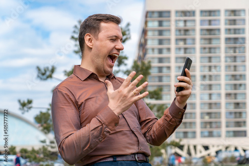 Fotografia, Obraz Portrait of handsome millennial man in a copper-colored shirt who received amazing news on phone, looking at smartphone screen, showing yes gesture and screaming yeah