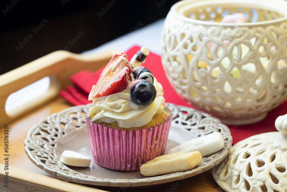 Pretty cupcake decorated with cream, strawberry, and berry fruits on openwork plate and wooden tray. Elegant dessert beautifully served.