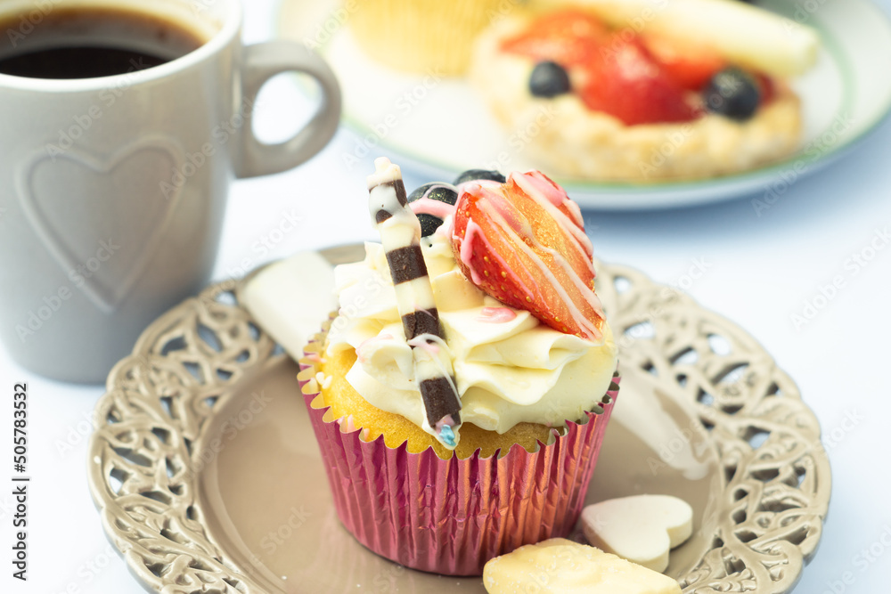 Whipped cream cupcake decorated with fruits, chocolate and gold sprinkles on romantic plate. Dessert and coffee in heart shaped ornament and other pastry.