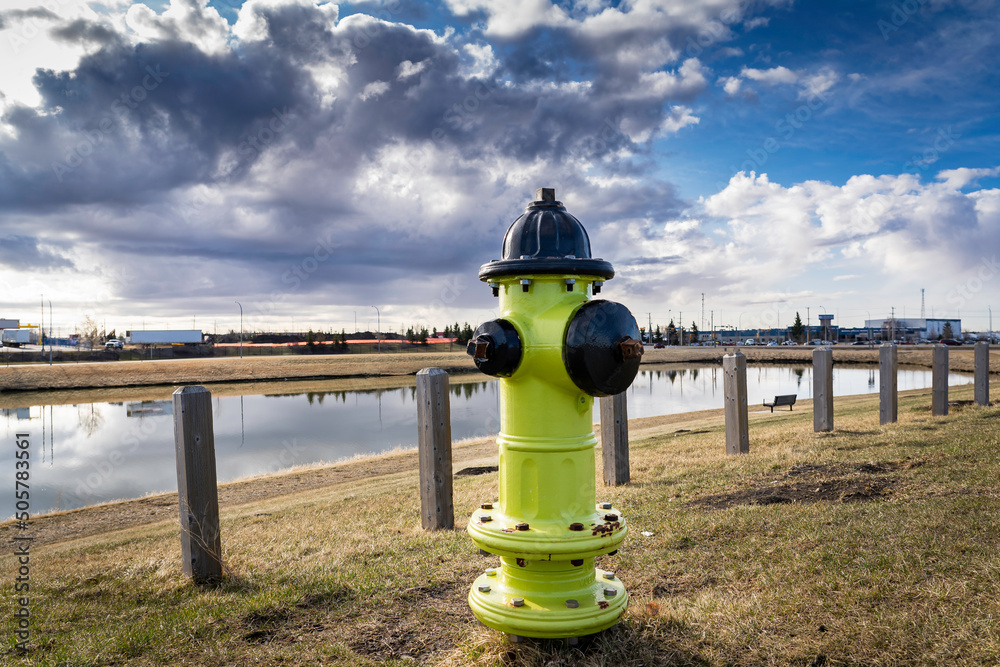 A yellow fire hydrant connected to infrastructure standing next to a storm retention pond in an industrial park in Airdrie Alberta Canada under a dramatic sky.