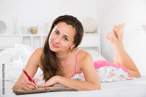 Portrait of young smiling woman lounging in bed and writing to diary in home interior photo