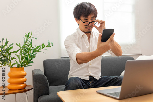 Middle age Asian man squint eye to look at the phone due to long eye sighted problems, which makes vision difficult.