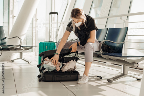 Portrait of woman wearing black T-shirt, white disposable mask, sitting on chair, packing clothes in black suitcase.