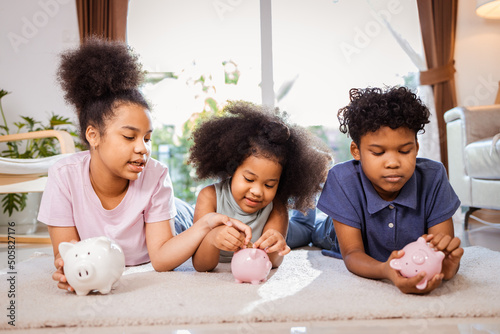 Photographie African American kids putting a coin into a piggy bank in a living room at home