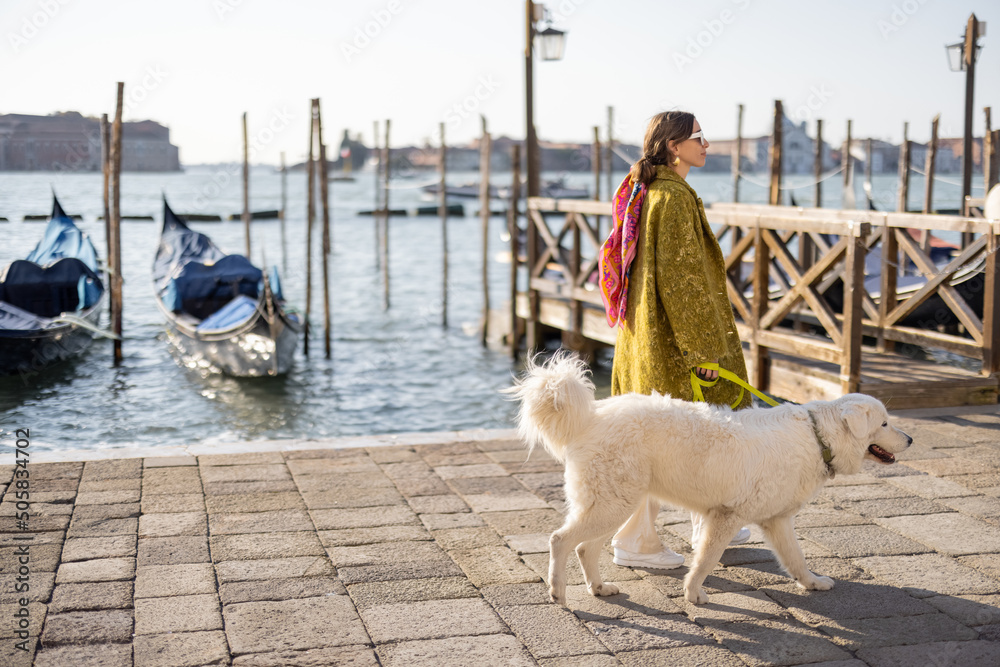 Young woman walking with dog near water canal with gondolas at saint Marks square in Venice. Woman wearing coat and colorful shawl in italian style. Maremmano abruzzese Italian sheepdog - obrazy, fototapety, plakaty 