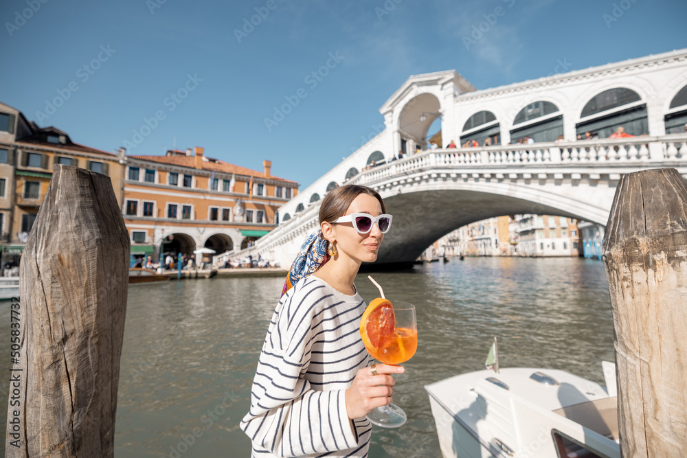 Young woman drinks summer cocktail on the background of famous Rialto bridge in Venice. Concept of happy vacation and leisure time in Italy. Holding italian alcohol drink spritz Aperol