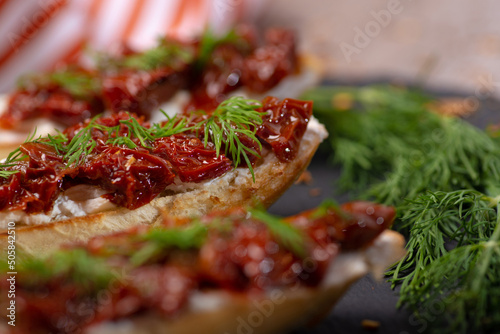 Bruschetta with the creamy cheese, dried tomatoes and the dill on the black stone plate on a blurred background, close up