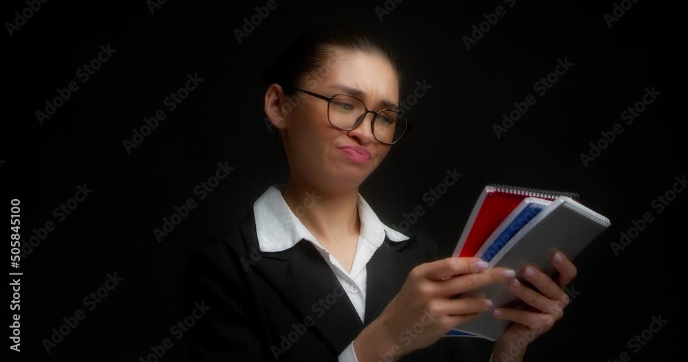 Brunette with glasses and business clothes does not like colorful notebooks in her hands. A business woman cannot decide on the choice of gray, blue or red. Isolated on a black background. Brunette with glasses and business clothes does not like colorful notebooks in her hands. A business woman cannot decide on the choice of gray, blue or red. Isolated on a black background.