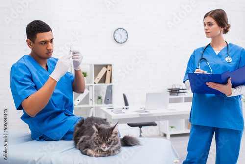 African american veterinarian holding syringe near blurred maine coon and colleague with clipboard in clinic