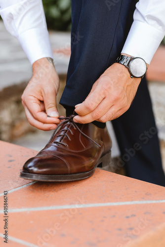 A businessman or groom is dressed up with classic elegant shoes in a outdoor space. Closeup view of male hands lacing beautiful elegant shoes on the wedding day