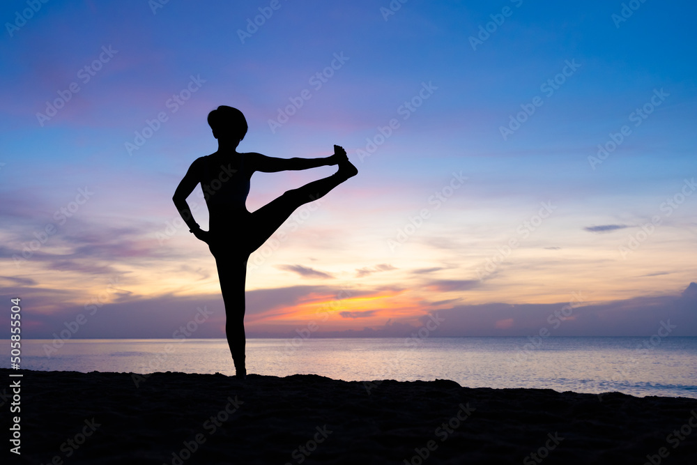 Silhouette of beautiful woman practicing yoga asana on the beach with sunrise on twilight blue vibrant sky and calm sea in background. Standing balancing, Stretching, Utthita Hasta, Challenging.