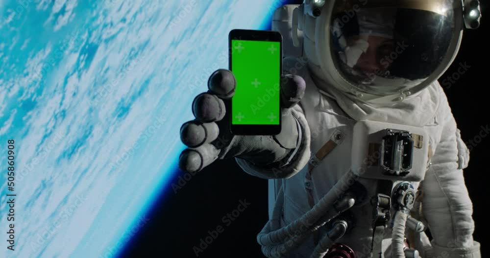 CHROMA KEY Caucasian female astronaut using her mobile phone during spacewalk near planet Earth, showing green screen into camera. Shot with 2x anamorphic lens