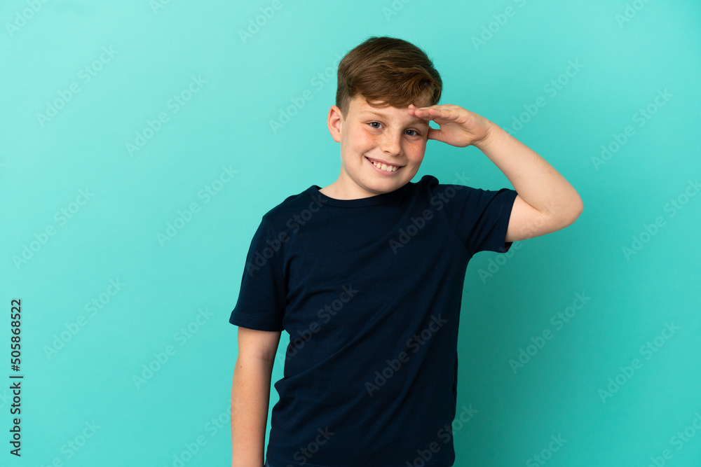 Little redhead boy isolated on blue background saluting with hand with happy expression Little redhead boy isolated on blue background saluting with hand with happy expression