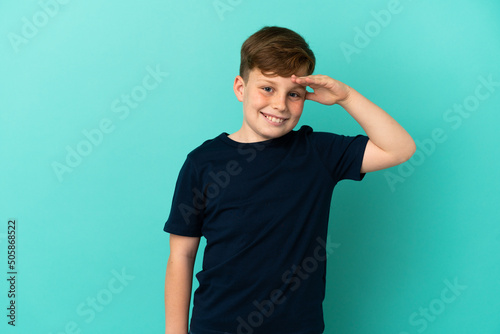 Little redhead boy isolated on blue background saluting with hand with happy expression