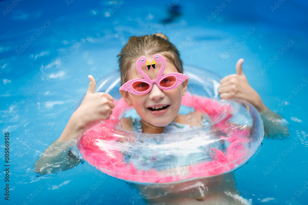 european preteen girl in flamingo sunglasses and a swimsuit swims on circle in pool in backyard. Funny child in pink glasses with shells and with an inflatable ring with pink feathers