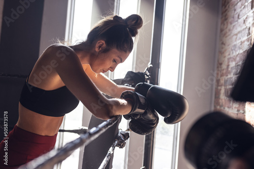 Side view of exhausted caucasian young female boxer wearing gloves standing by ropes in boxing ring photo