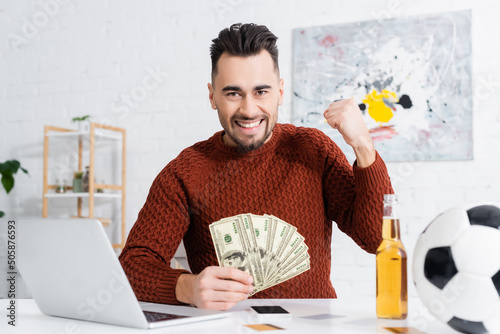 excited bookmaker with dollar banknotes showing win gesture near laptop and soccer ball. photo
