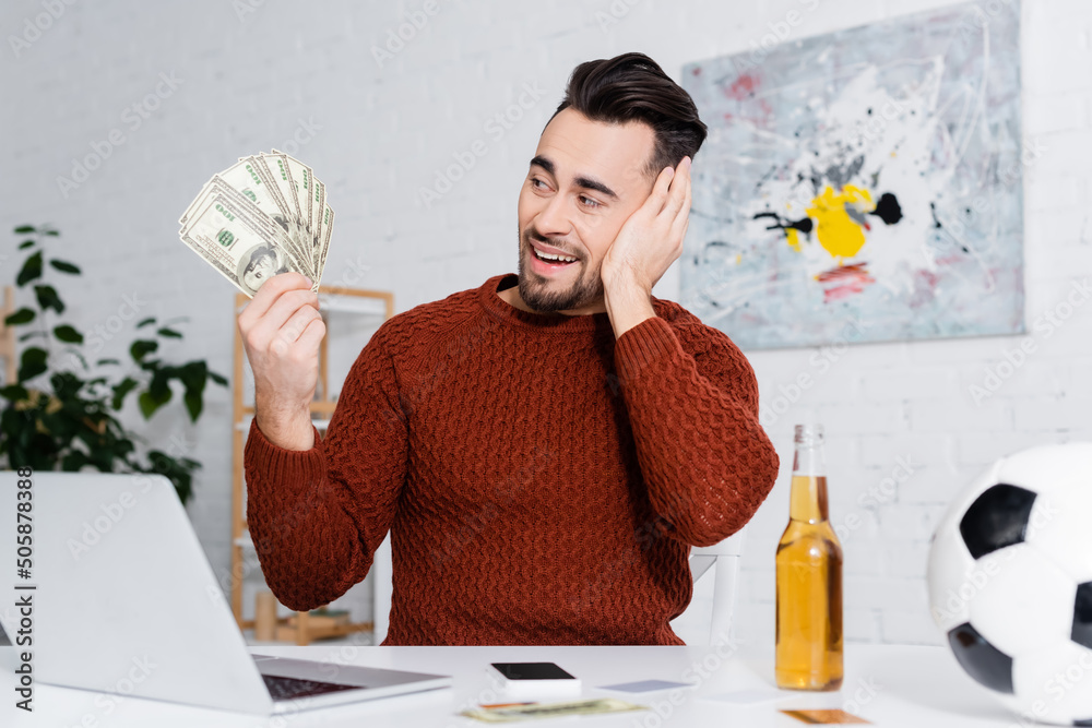 Fototapeta amazed bookmaker holding dollars near laptop, bottle of beer and soccer ball
