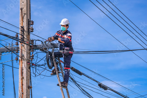 Asian technician in protective mask on wooden ladder is installing internet fiber optic line on electric power pole against blue sky background