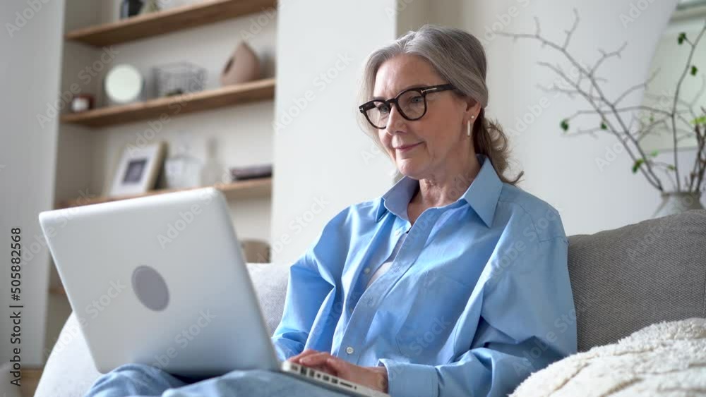 Senior female typing email on laptop, blogging, writing message, working on project from home while sitting on the couch in the living room Senior female typing email on laptop, blogging, writing message, working on project from home while sitting on the couch in the living room