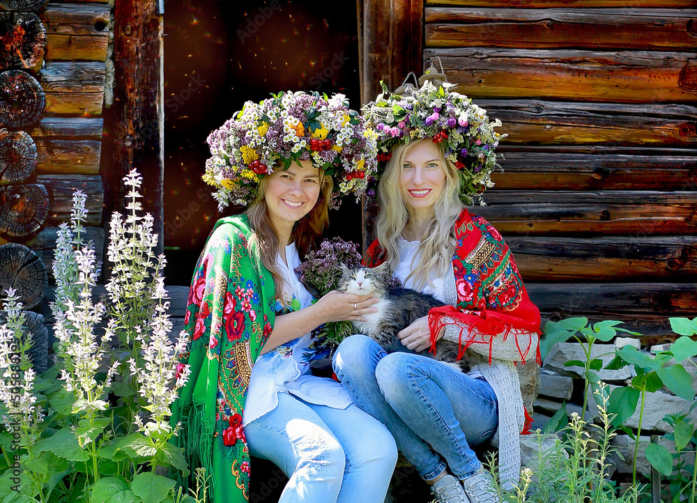 Two girls in large flower wreaths and national scarves on their shoulders are sitting with a cat. Background rural wooden house. Nature. Summer concept.