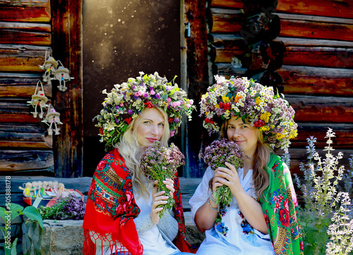 Young girls in large flower wreaths, Ukrainian national scarves on their shoulders with bouquets sitting. Background rural wooden house. Traditions in Ukraine. Summer concept.