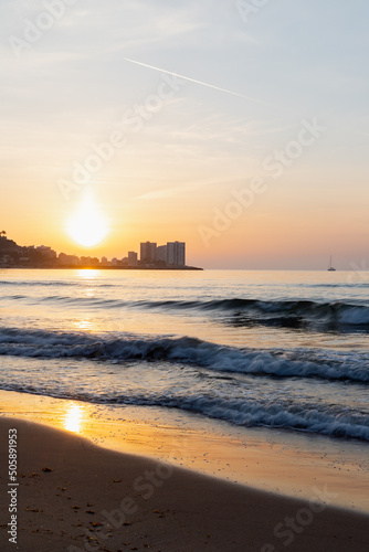 sunrise on the beach, sun rising behind the mountain, small boat in the background, waves of the sea, plane at top, vertical image