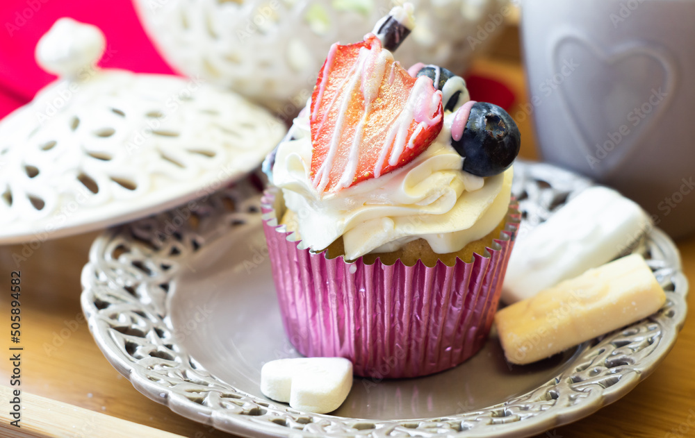 Romantic cupcake decorated with cream, strawberry, and berry fruits on a Victorian openwork plate and wooden tray. Elegant dessert beautifully served among coffee mug and old vintage porcelain.