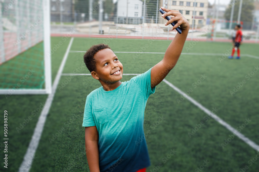 Kid using phone. Smiling and excited boy showing smartphone empty screen. Looking to camera. Cellphone display mockup mobile app advertisement. African american kid in football field. Blank screen Kid using phone. Smiling and excited boy showing smartphone empty screen. Looking to camera. Cellphone display mockup mobile app advertisement. African american kid in football field. Blank screen