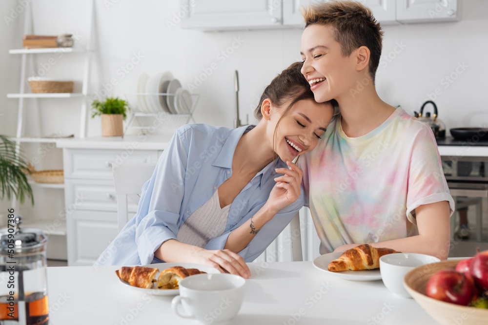excited pansexual partners laughing in kitchen near croissants and tea cups.