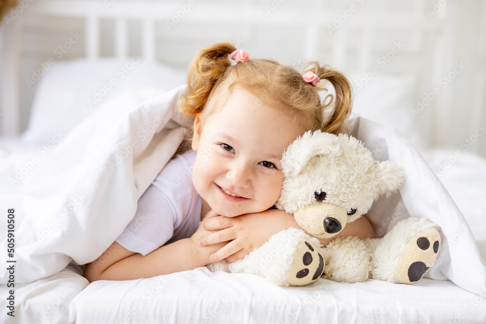 a little cute baby girl peeks out from under a blanket on a white cotton bed at home smiling hugging a teddy bear
