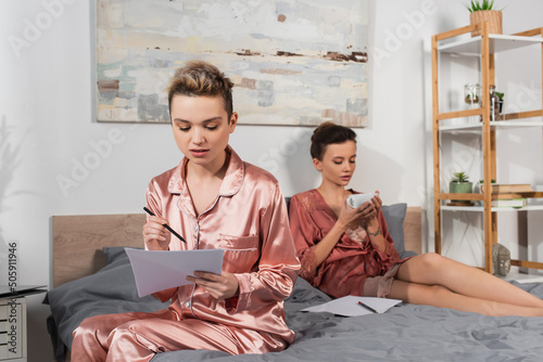 pangender person in satin pajamas working with papers near partner drinking tea on bed. photo