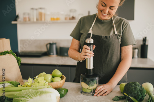 Young woman making smoothie at the kitchen, using blender and fresh fruits and vegetables.