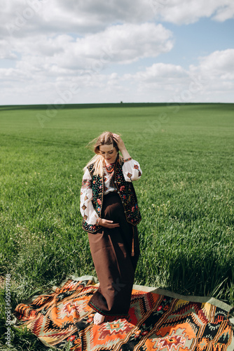 Young pregnant Ukrainian woman in ethnic national dress on a wheat field on an embroidered antique carpet