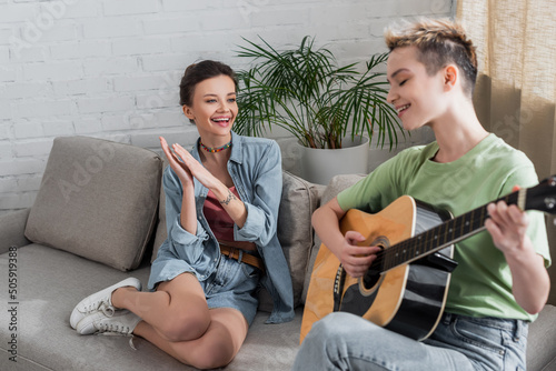 pleased pangender person applauding to lover playing guitar in living room. photo