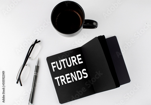 FUTURE TRENDS written text in small black notebook with coffee , pen and glasess on white background. Black-white style