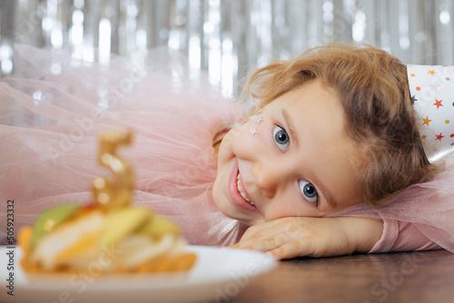 Children's positive emotions. Beautiful portrait of charming girl looking into camera, who put her head on palm of her hand located on table and is satisfied. Front cake with number five.