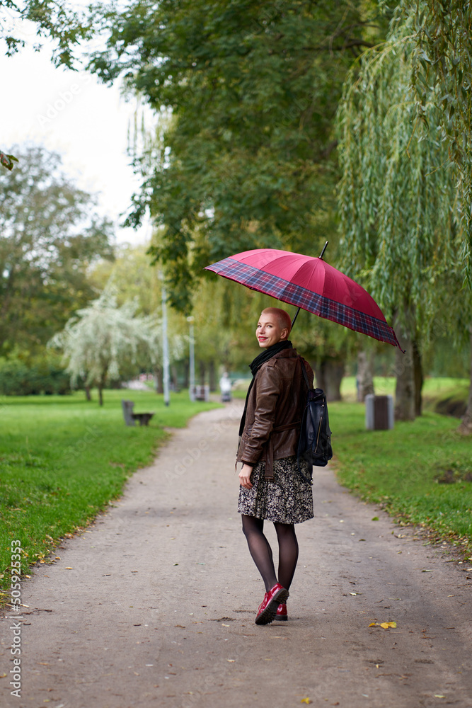 Young smilling short haired blonde woman walks in the autumn park with red umbrella. View from back. Young smilling short haired blonde woman walks in the autumn park with red umbrella. View from back.