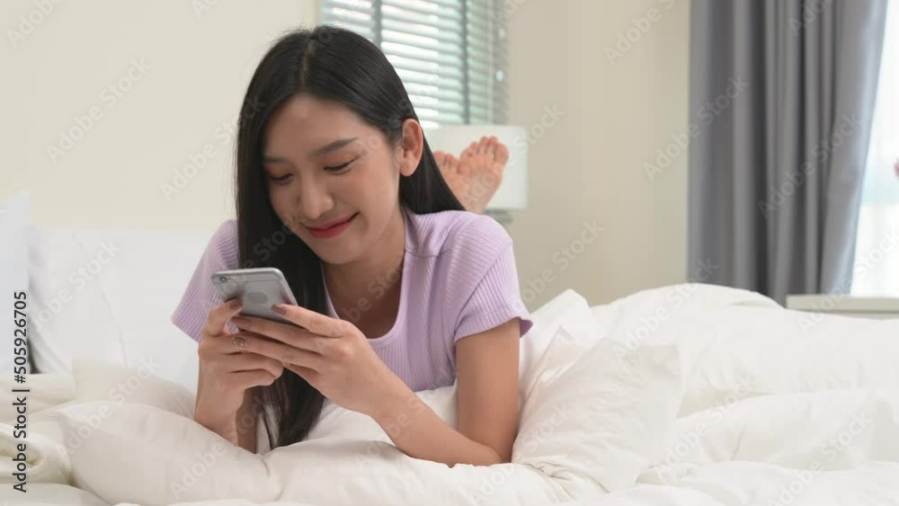 Young asian lady lying on the bed in her bedroom playing with her mobile phone and chatting, texting, messaging with her friends in a happy and cheerful mood and feels.